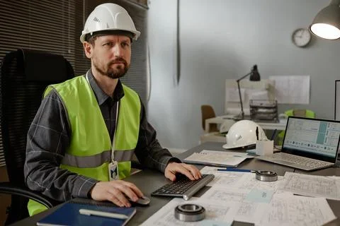 Bearded mechanical engineer wearing hardhat using computer at workplace Stock Photos