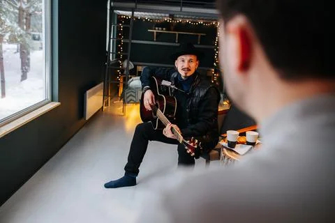Bearded middle aged man in hat sitting with guitar, looking at man in foregro Foto stock