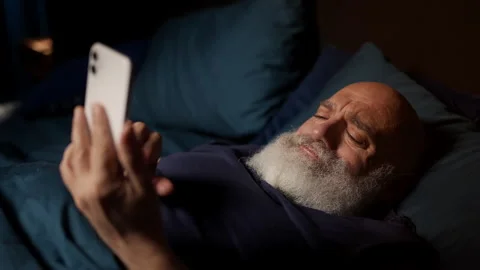Bearded middle-aged man reads hurtful texts on phone in bed, feeling depressed Stock Footage 342520889