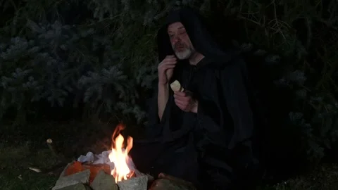 A bearded old monk eating bread, eating supper at night by a fire in the woods. Stock Footage 303783302