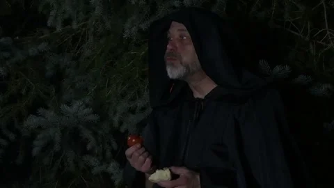 A bearded old monk eats an apple and a tomato at night by a fire in the woods. Stock Footage 303783286