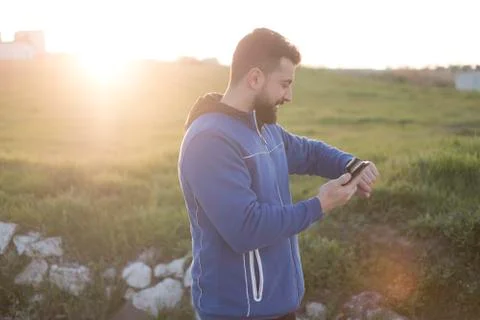 Bearded runner resting after training outdoors, comparing phone to smart watc Stock Photos