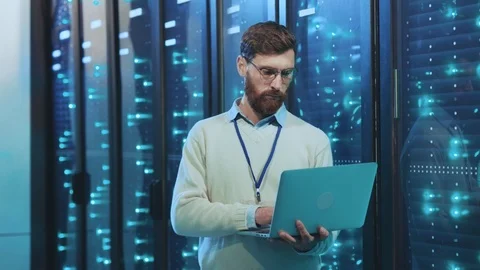 Bearded smart IT specialist working on laptop standing at operational rack Stock Footage 119233168