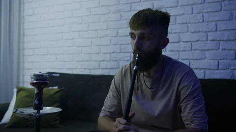 A bearded stylish man is sitting on the couch at home and smoking a hookah Stock Footage 204843910