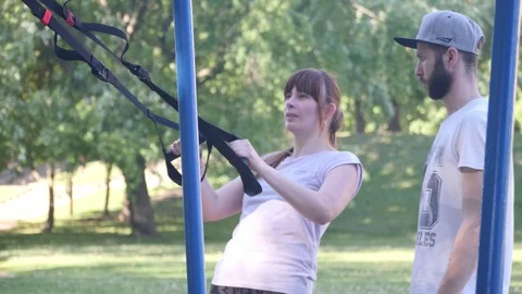 Bearded trainer in baseball cap controlling making pull-ups on fitness loops by Stock Footage 98568939