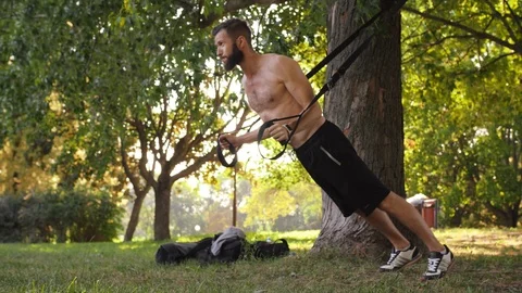Bearded young athlete making push-ups on fitness loops near a tree in the park Video stock 99162642