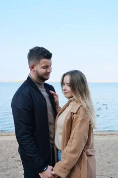 A bearded young man in a black jacket looks at a girl in a coat and holds her Stock Photos