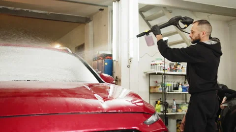 Bearded young man car wash worker spraying cleaning foam to a modern red car Stock Footage 233997649