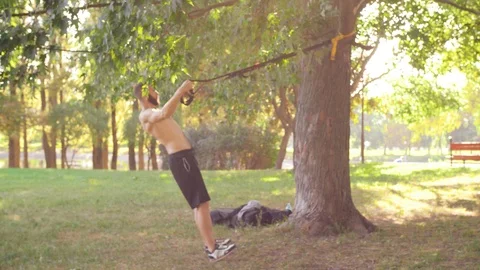Bearded young man doing pull-ups on fitness loops in a sunshine park. 4K slow mo Stock Footage 99161968