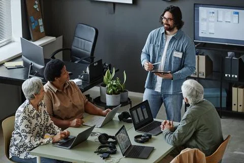 Bearded young man leading computer class for seniors using laptops at table Stock Photos