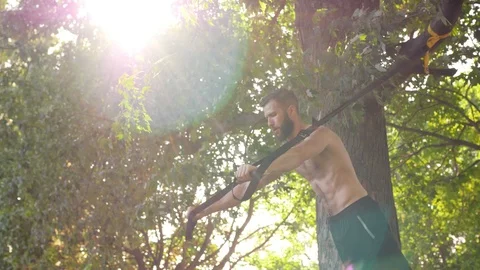 Bearded young man making push-ups on fitness loops near a tree in sunshine park Stock Footage 99201392