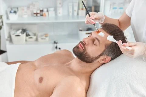 Bearded young man is relaxing while cosmetologist is spreading white clay on his Stock Photos