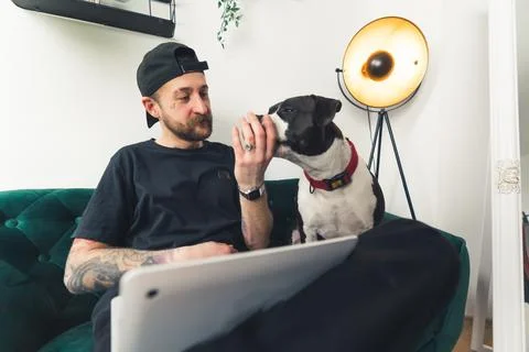 A bearded young man with tattoos cap and laptop cuddling his dog medium full Stock Photos