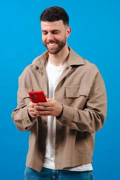 Bearded young man texting on red smartphone, wearing jacket on a blue background Stock Photos