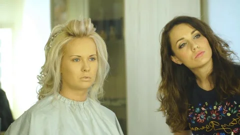 Beautiful blonde considering the makeup in the mirror with the makeup artist Stock Footage 76940005