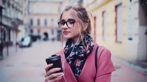 Beautiful blonde girl with headphones sips coffee in the city street Stock Footage