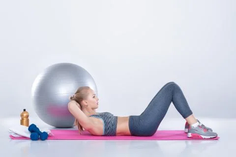Beautiful blonde girl in sportswear does exercises on fitness Mat on gray Stock Photos