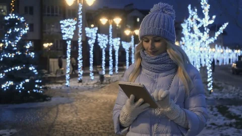 Beautiful blonde in winter clothes uses a tablet on the night lights background Stock Footage