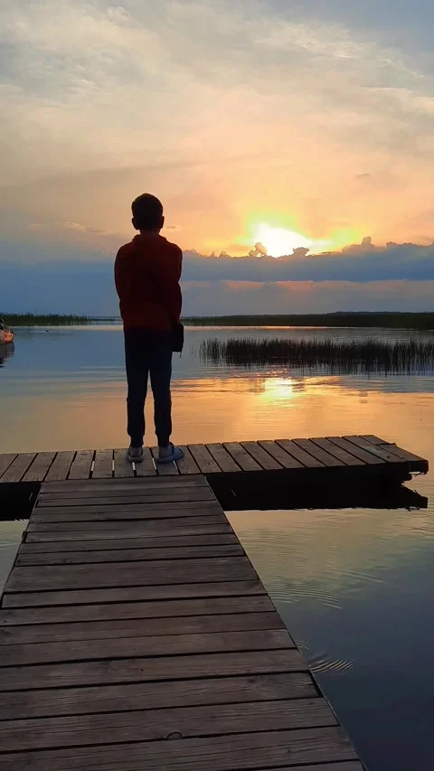 A beautiful boy standing on a dock watching the sun set,  lake Stock Footage 280892195