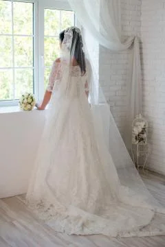 Beautiful bride looking out the window.In the white room back view. in full.. Stock Photos