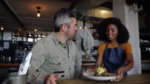 Beautiful ethnic waitress serving middle-aged male egg breakfast at trendy cafe. Stock Footage 138592381