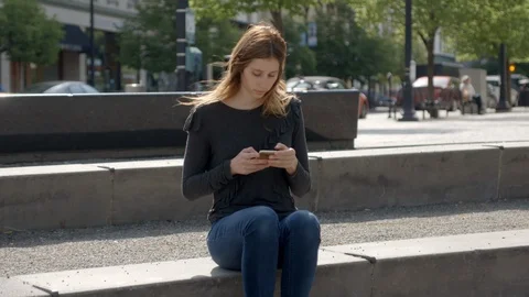 Beautiful female using cellphone on bench downtown with hair gently blowing Stock Footage 90023885