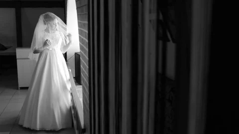Beautiful, gentle bride standing at the window. Adjusts her veil. Looking out th Vidéo 73636888