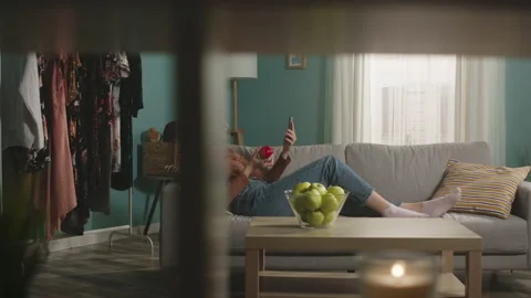 Beautiful girl eats a red apple, lying on sofa and and speaks via video link Stock Footage 151963142