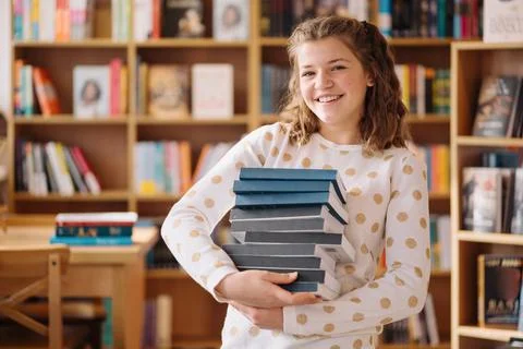 Beautiful girl is holding stack of books while standing among books in the Stock Photos