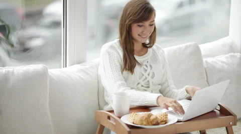 Beautiful girl sitting by window using laptop drinking from cup and smiling  Video stock 34150108