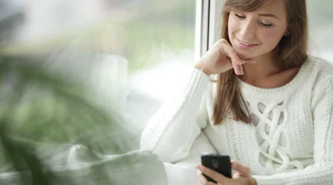 Beautiful girl sitting by window using mobile phone looking at camera  Video stock 34150118
