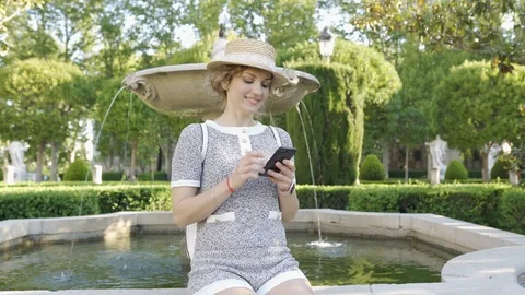 Beautiful girl tourist in a hat in a beautiful garden with a fountain is looking Stock Footage