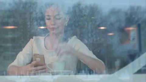 Beautiful girl using phone while drinking coffee in a cafe viewed through window Stock-Footage 71497389