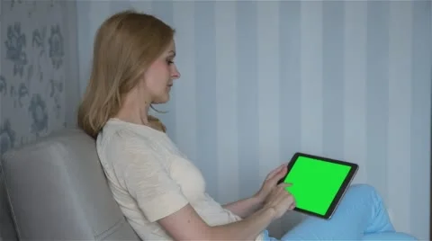 Beautiful girl using tablet pc with pre-keyed green screen sitting on sofa Stock Footage 63524649
