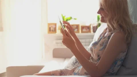 Beautiful girl using Tablet PC sitting on sofa near window 2 sun glare Stock Footage 65220018