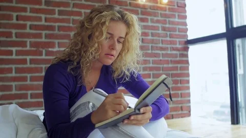 Beautiful girl writes note in a notebook sitting on her bed and smiling close up Video stock 83597366