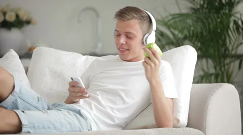 Beautiful guy relaxing on sofa using cellphone listenning music and eating  Stock Footage 27692068