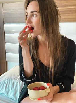 Beautiful lady in hotel bed eats strawberry in black pajamas and looking away Stock Photos