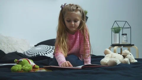 Beautiful little girl sitting on the bed and reading a book at home Stock Footage