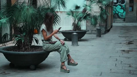 Beautiful long-haired brunette texting message while sitting on a street pot Stock Footage 139442432