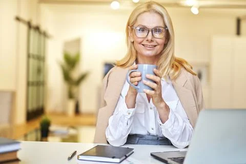 Beautiful mature businesswoman in glasses smiling at camera and drinking coffee Stock Photos