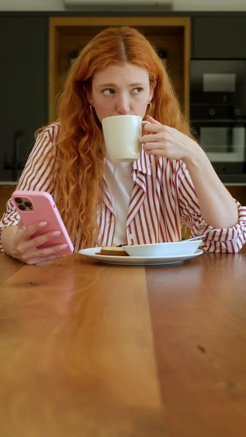 Beautiful redhead woman in pajamas using her pink smartphone and drinking Stock-Footage 344641925