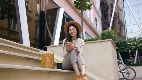 Beautiful slender smiling young lady in modern clothes and trendy hat sitting on Stock Footage 120185012