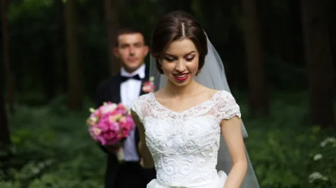 Beautiful smiling bride goes forward through the green park while groom with Stock Footage 64032137