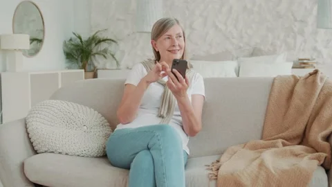 Beautiful smiling elder middle age woman using smartphone sitting on sofa at Vídeos de archivo 224538916
