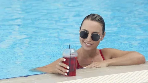 Beautiful smiling girl is resting by the pool in sunglasses, drinking coctail Stock Footage