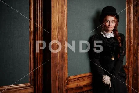 Beautiful Victorian woman in black dress and top hat stands in vintage interi Stock Photos