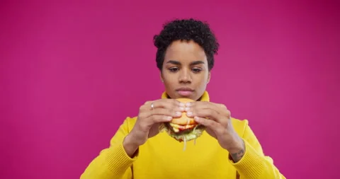 Beautiful woman with afro eating burger on pink background enjoying delicious Stock Footage
