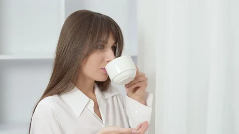 A beautiful woman is drinking hot tea from a cup standing by the window Stock Footage 166310894