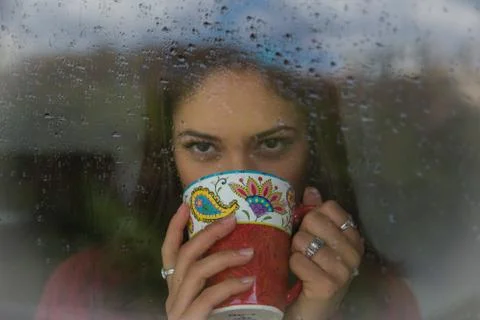 Beautiful woman looking through the window and drinking tea on a rainy day. Stock Photos
